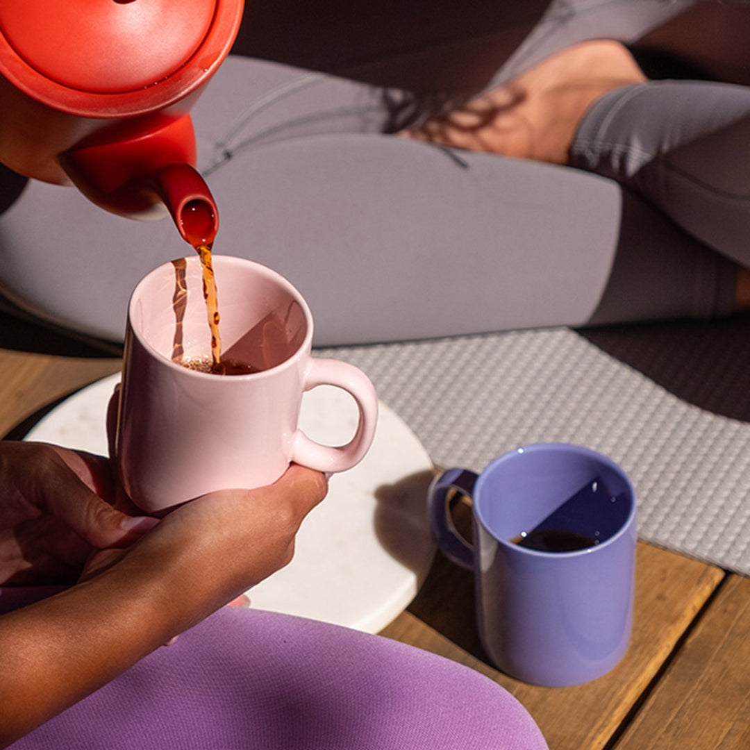 Person pouring tea from a red teapot into a pink mug on a wooden table.